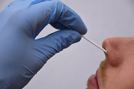 Hand im blauen Einweghandschuhe steckt Corona-Teststäbchen in die Nase eines Mannes